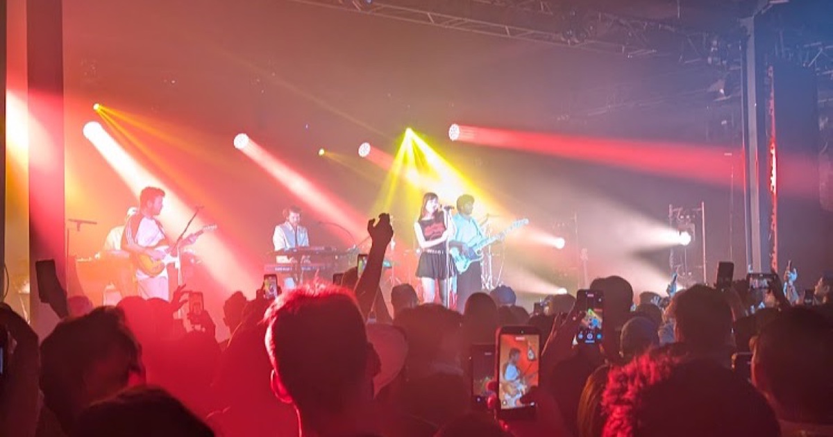A Bangkok livehouse mid-show, crowd silhouettes and phones up under red and yellow stage lights